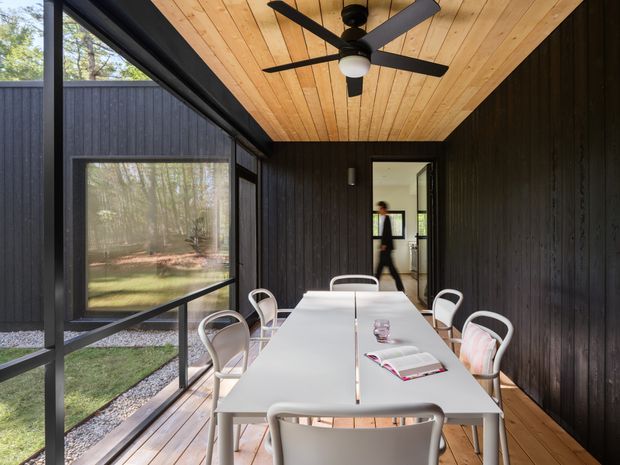 A covered outdoor dining terrace enclosed by dark vertical timber-clad walls and large glazed panels. A long white dining table with white chairs occupies the space, while the ceiling is lined with warm timber boards and fitted with a black ceiling fan. The terrace overlooks a landscaped garden and connects directly to an interior room through an open doorway.