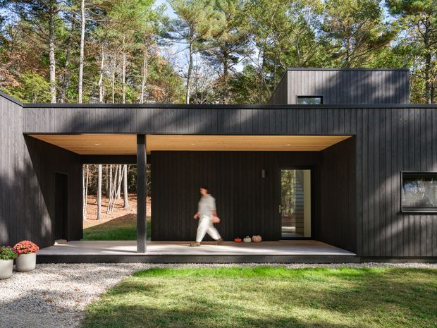 Exterior view of a contemporary single-storey dark-clad house set among mature trees. The facade features vertical black cladding, a strongly recessed covered entry or outdoor passage with a warm timber soffit, simple rectangular glazing, and a flat-roofed composition. Gravel and lawn occupy the foreground, while the sheltered threshold creates a strong indoor-outdoor connection.