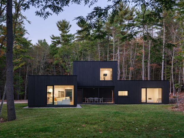 Exterior view of a low, modern black-clad house set within a dense wooded landscape. The residence is composed of offset rectangular volumes with a taller central upper box, large warm-lit windows, and a recessed covered outdoor dining or entry area. The dark facade contrasts with the surrounding trees and lawn, creating a minimalist contemporary architectural composition.