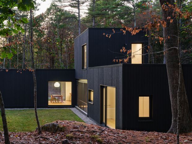 Exterior view of a compact contemporary house composed of offset dark-clad volumes set within a wooded site. The black vertical cladding contrasts with warm illuminated windows and glazed openings, while a small lawn, exposed rock and mature trees surround the building.