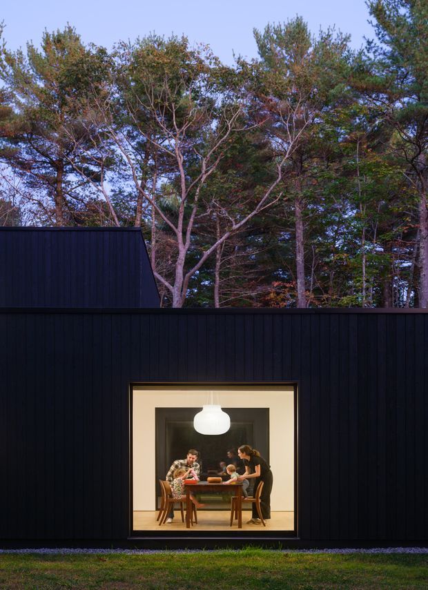Exterior view of a compact, dark-clad modern building set among mature trees. A large square window reveals a warmly lit dining room with people gathered around a dining table, while a simple pendant light hangs prominently inside. The composition contrasts the near-black vertical cladding with the surrounding woodland and evening-blue sky.
