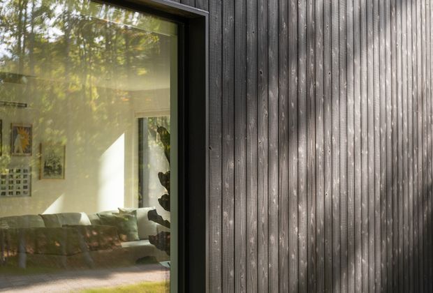 Close-up exterior view of a contemporary building facade clad in vertically arranged dark timber boards with a black-framed glazed window. The window reflects surrounding vegetation and reveals part of a softly furnished living room inside.