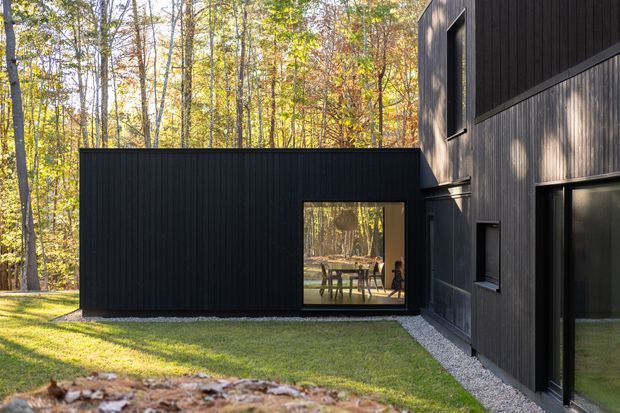 Exterior view of a dark-clad contemporary house set within a wooded landscape. The composition shows a low rectangular wing connected to a taller volume, with vertical dark cladding, large glazed openings, and a gravel edge bordering the lawn. Warm sunlight and autumn trees are visible around the building.