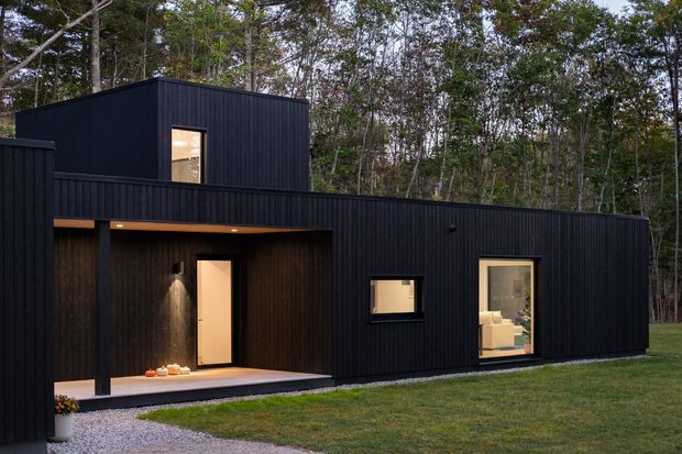 A contemporary single-storey black-clad house set within a wooded site. The exterior features vertically ribbed dark cladding, a projecting upper volume, warm timber-lined recessed entry, and large rectangular windows with warm interior light visible through them. A gravel path and lawn lead to the entrance.