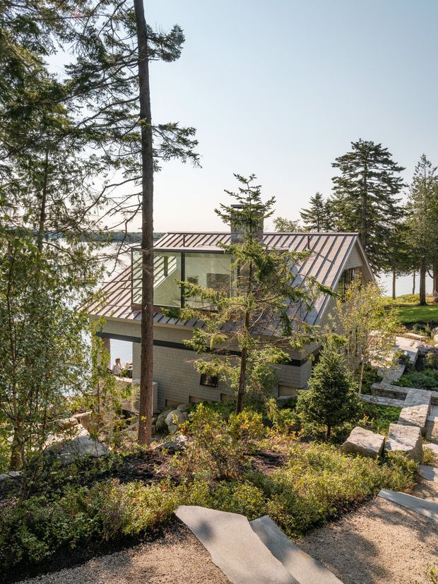 Exterior view of a contemporary house set among mature trees on a landscaped, sloping site. The building has a steeply pitched metal roof with prominent gable forms, large glazed openings, and light grey masonry or cladding. Stone steps and paths weave through naturalistic planting, with a bright body of water visible beyond the house and trees.