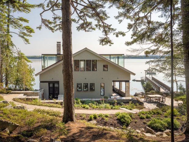 Exterior view of a contemporary gable-roofed waterfront home set among mature trees and landscaped grounds. The house has light grey horizontal cladding, extensive glazing including a glazed upper-level section, dark-framed windows and doors, and a covered outdoor area facing the water. A timber dock and stair access lead down to the shoreline.