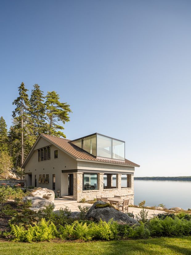 Exterior view of a two-storey lakeside house set among exposed granite rocks, mature conifers, and ferns. The home features a gabled standing-seam metal roof, light horizontal siding, a stone-clad lower level, large glazed openings, and a contemporary glass rooftop volume. A covered terrace with outdoor dining furniture faces the water.