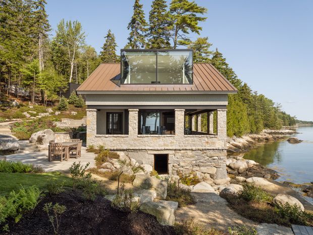 Exterior view of a compact stone-clad lakeside or waterfront house set into a rocky, landscaped site. The building has a steeply pitched copper-toned metal roof, dark-framed windows, a projecting glazed upper-level section, and a covered outdoor dining terrace beside the water. Natural stone walls, large boulders, mature trees, and shoreline landscaping dominate the setting.