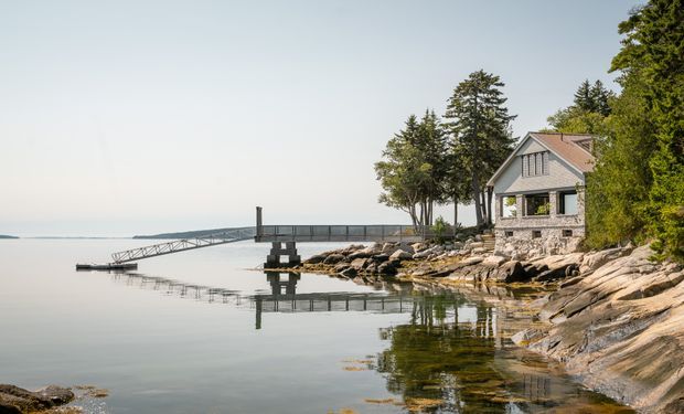 A tranquil waterfront exterior showing a stone-clad, gabled house set among exposed rocky shoreline and mature evergreen trees. A long dock extends across calm reflective water, and the house features a pitched roof, timber-framed gable detailing, and large glazed openings.