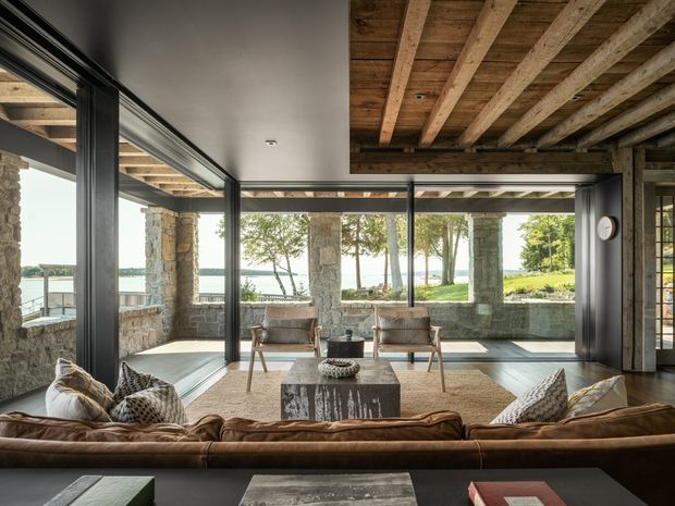 A spacious, rustic-contemporary living room opening onto a stone-covered outdoor terrace and waterfront landscape. The room features expansive black-framed glazing, a warm brown leather sectional sofa, lounge chairs, a stone coffee table, and a striking exposed timber-beam ceiling. Natural stone columns and walls continue through the indoor-outdoor connection, creating a substantial, lodge-like architectural character.