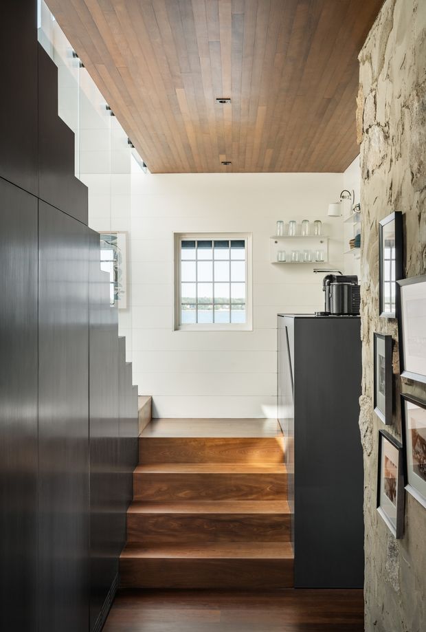 A compact, refined interior stairway leading up to a small coffee or kitchenette area. The staircase has rich dark timber treads and is enclosed by sleek matte-black built-in cabinetry on the left. A rough exposed stone wall with framed artwork lines the right side, while white horizontal wall panelling, a gridded window, open glass shelving, and a black coffee cabinet occupy the upper level. The ceiling is clad in warm timber boards with recessed downlights, creating a strong contrast between warm wood, black joinery, white walls, and textured stone.