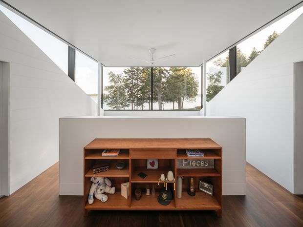 A bright, minimalist interior with a low central timber display cabinet positioned in front of a half-height white partition. Dark timber flooring, white angled walls and ceiling, black-framed floor-to-ceiling glazing, and a ceiling fan create a clean contemporary architectural setting. Trees are visible outside through the glazing.