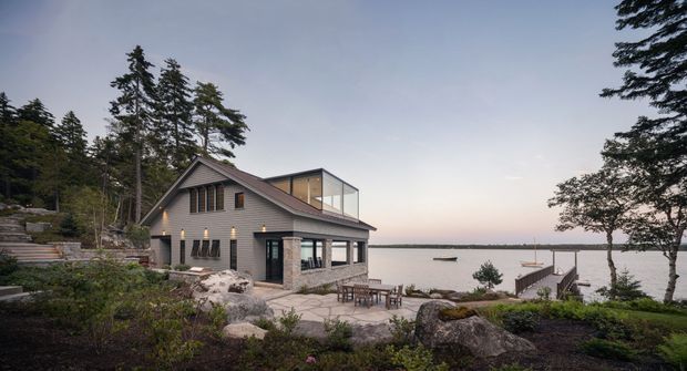 A contemporary waterfront residence set among rocky landscaped terrain and tall trees. The house features a prominent pitched gable roof, dark grey exterior walls, timber-toned vertical cladding accents, and expansive glazing. A paved terrace with outdoor dining furniture leads down toward a small private dock and calm water beyond.