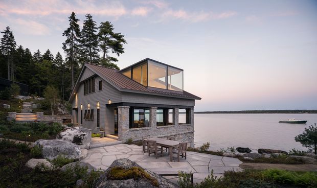 A contemporary lakeside house viewed from the landscaped waterfront, featuring grey horizontal cladding, a dark pitched metal roof, stone masonry at the lower walls, and a glazed rooftop-style clerestory volume. A paved terrace with outdoor dining overlooks the calm water, while exposed bedrock and mature conifers reinforce the natural setting.