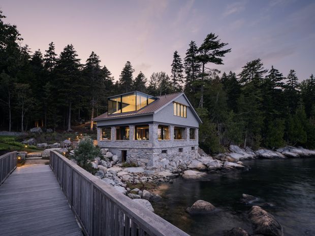 A contemporary two-storey coastal house set among dense forest and exposed rocky shoreline, viewed from a timber pedestrian walkway over the water. The home has a stone-clad lower level, horizontal light-grey weatherboard cladding above, a pitched metal roof, extensive warm-lit glazing, and a projecting glazed upper volume. Rocks, trees, and calm water surround the building at dusk.