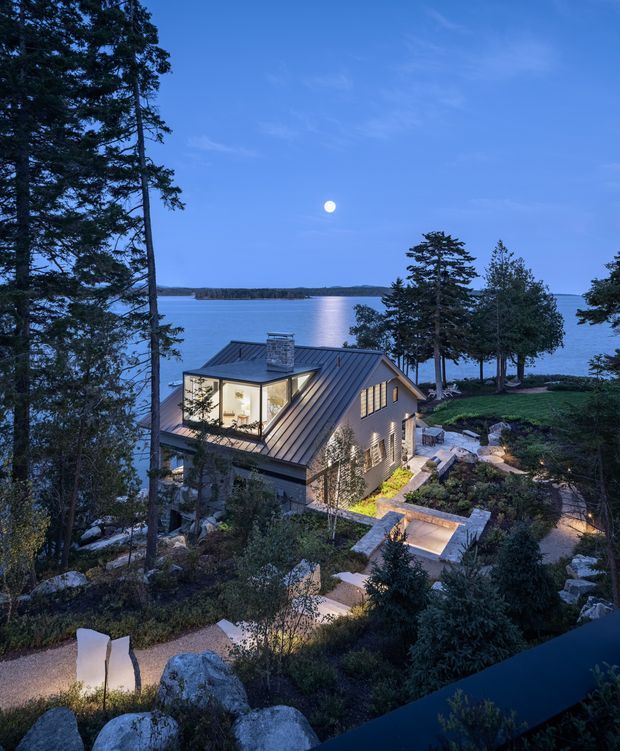 Twilight exterior view of a compact pitched-roof house set on a rocky, landscaped waterfront site. The home has dark standing-seam metal roofing, extensive illuminated glazing, stone-clad walls, and a chimney. Terraced paths, retaining walls, steps, and outdoor seating areas descend toward a calm body of water, with tall trees framing the scene and a moonlit blue sky beyond.