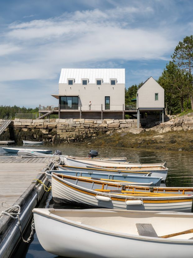 Exterior view of a contemporary waterfront residence set above a rocky shoreline, photographed from a dock with several small boats in the foreground. The main house has a simple rectangular form, pale horizontal cladding, a prominent white standing-seam metal gable roof, large glazed doors, and a raised timber deck. A smaller matching gabled structure sits to the right on supports over the waterfront.