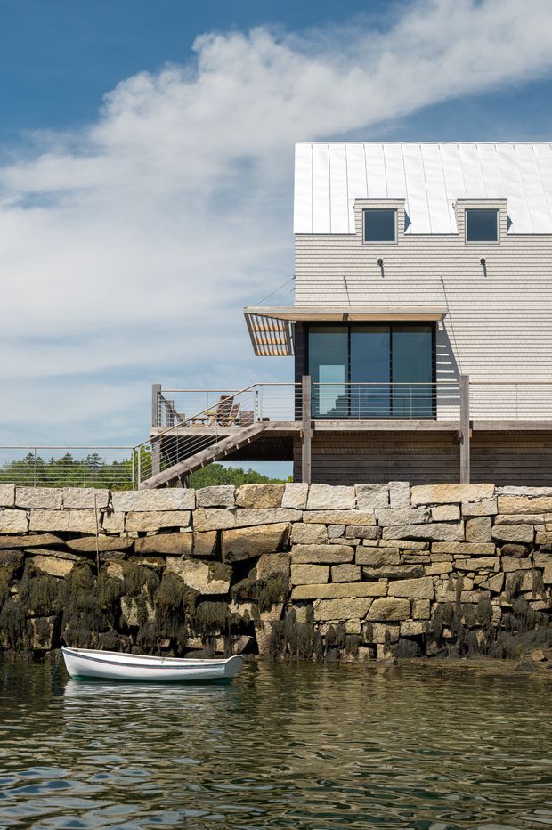 Exterior view of a contemporary coastal multi-level house beside the water. The building has a bright standing-seam metal roof, pale horizontal cladding, dark-framed glazing, a covered balcony or deck, and an exterior stair descending toward a substantial rough-stone seawall. Water and a small boat are visible in the foreground.