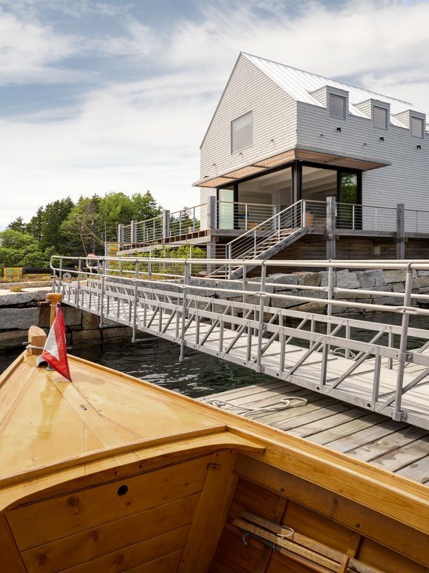 Exterior view of a contemporary multi-storey waterfront house with a steep gabled roof, pale horizontal cladding, dark-framed glazing, and a timber deck with metal railings. A gangway leads down to the water, seen from the bow of a wooden boat in the foreground. The setting includes rocky shoreline landscaping and mature trees.