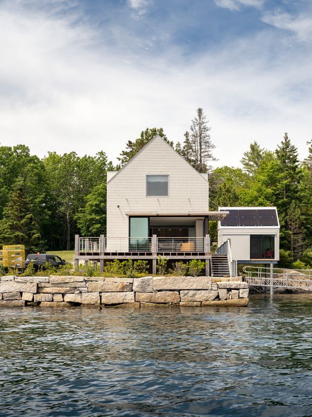 Exterior view of a contemporary waterfront residence photographed from across the water. The house has a steep front gable, pale horizontal cladding, large glazed openings, elevated timber decks and balconies, a smaller adjoining wing with rooftop solar panels, and a substantial stone shoreline retaining wall. Dense trees surround the property.