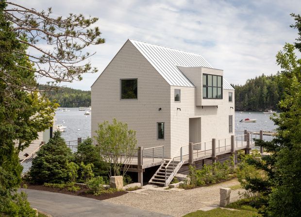 Exterior view of a compact contemporary waterside house set among mature trees. The home has pale horizontal cladding, a steep gabled metal roof, projecting upper-level glazing, and raised timber decking with stairs leading to a gravel path. A sheltered inlet and wooded shoreline are visible behind the building.