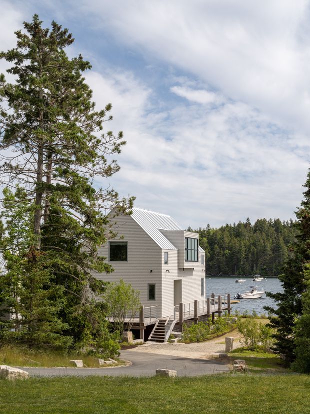 A contemporary two-storey lakeside house clad in pale horizontal siding, with a steep standing-seam metal gable roof, large glazed upper-level projection, and timber waterfront deck. The home sits among mature coniferous trees beside a small dock and calm lake, reached by a curved driveway and exterior stairs. The scene is bright and natural with a strong forest and waterfront setting.