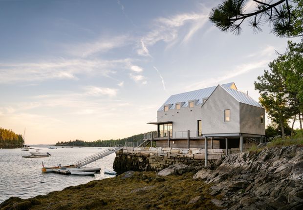 A contemporary coastal house perched on a rocky shoreline beside calm water. The elevated structure has pale grey horizontal cladding, a steep metal roof with multiple skylights and dormer-like roof windows, extensive glazing, and a timber deck overlooking the waterfront. Pine trees and distant wooded shoreline frame the scene.