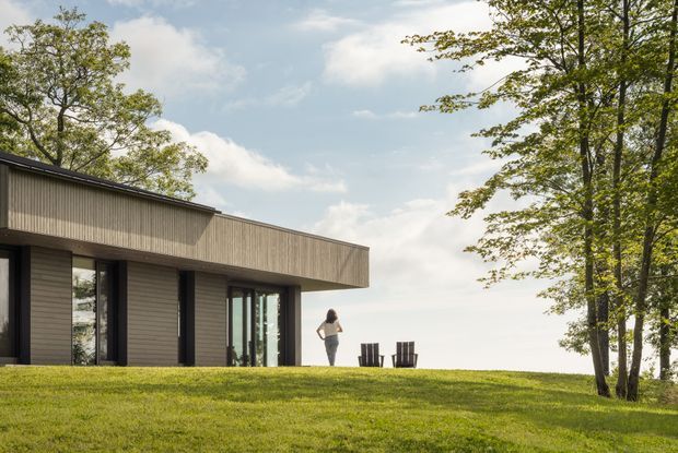 A low, modern single-storey residence set in a gently landscaped grassy clearing, with a broad flat roof and deep overhang. The facade combines dark horizontal wall cladding with vertically ribbed upper cladding, large dark-framed glazed openings, and a glazed corner or sliding door. A person and outdoor chairs provide scale, while mature deciduous trees surround the house.