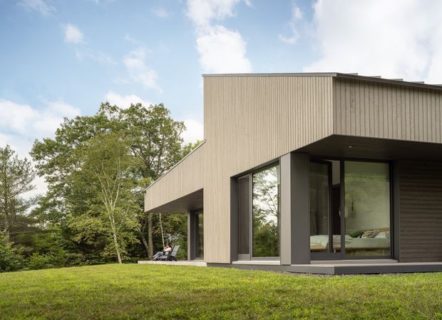 A contemporary single-storey house set within a landscaped, wooded site. The building has a strong rectilinear form with a low-pitched or flat roof, vertical taupe-grey cladding, deep overhangs, and large dark-framed glazed openings. A bedroom is visible through the corner glazing, while the foreground shows a broad lawn and mature trees.