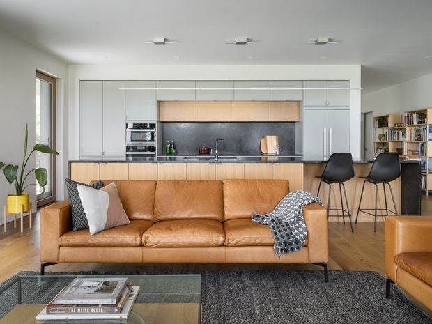 A bright, open-plan contemporary living area with a tan leather sofa in the foreground and a streamlined kitchen behind it. The kitchen features extensive pale grey built-in cabinetry, light timber overhead cupboards, a dark stone-look splashback, integrated appliances and a peninsula with black bar stools. Timber flooring, a large dark textured rug, a glass coffee table, open shelving and indoor plants complete the warm, modern interior.