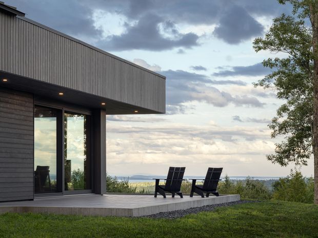 A modern single-storey house exterior with a deep cantilevered flat roof, dark vertical and horizontal cladding, and full-height glazed doors opening onto a raised timber deck. Two outdoor lounge chairs face a distant water view, with lawn and trees surrounding the home under a dramatic cloudy sky.
