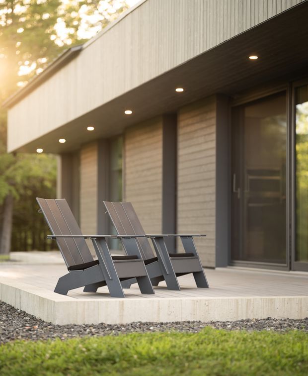 Exterior architectural photograph of a contemporary single-level building with a broad overhanging roof or fascia, recessed downlights, vertical light-coloured cladding, dark-framed glazing and a textured concrete or masonry wall. Two modern outdoor lounge chairs sit on a raised concrete patio, with a landscaped wooded setting around the building.