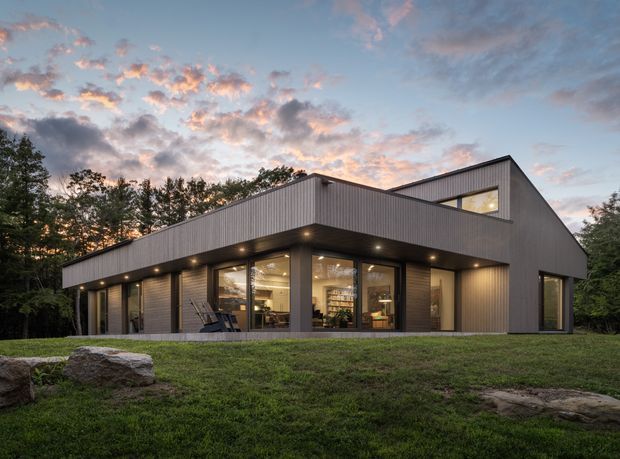 Exterior view of a contemporary single-storey house set in a wooded landscape at dusk. The home has a strongly articulated asymmetric gabled roof, dark vertically ribbed metal cladding, broad floor-to-ceiling glazed openings, deep roof overhangs with recessed downlights, and warm interior illumination. A small upper-level clerestory-like window is visible beneath the raised roof form.