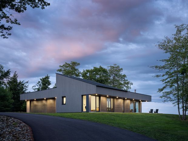 A modern single-storey house photographed at dusk, set on a landscaped sloping site. The low elongated building has dark vertical cladding, a sharply angled shed-style roof, warm recessed exterior lighting, large glazed openings, and an asphalt driveway curving around the front. Trees and lawn surround the home, with a bright open horizon visible beyond.