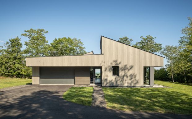 A single-storey contemporary house viewed from the front, set within a leafy landscaped site. The building has a distinctive asymmetrical steeply sloping roof form, light vertical wood-like cladding, dark horizontal cladding at the recessed garage and entry, and large dark-framed glazed openings. A paved driveway and path lead toward the house.
