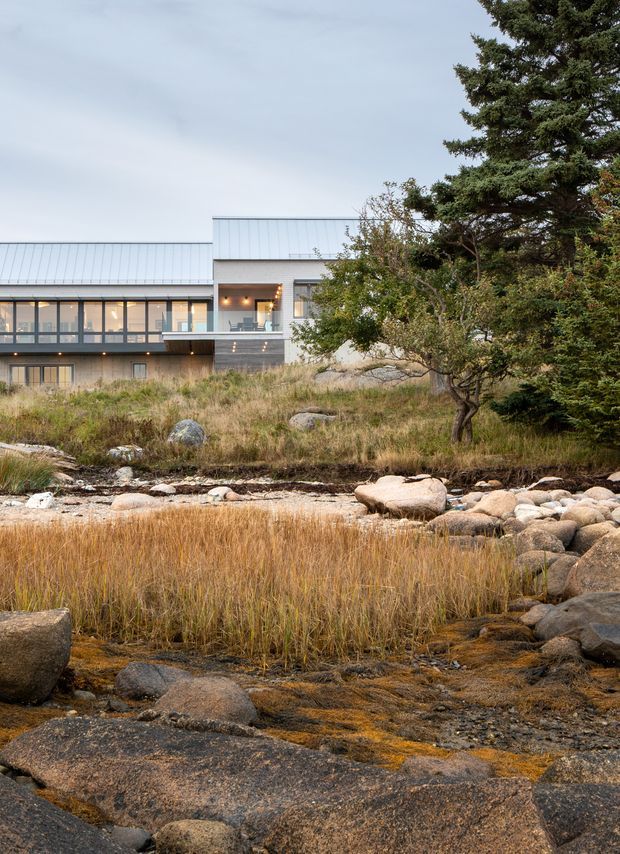 Exterior view of a contemporary multi-level house set within a rugged natural landscape. The building features a low-pitched metal roof, pale metal cladding, extensive dark-framed glazing, and projecting balcony or terrace areas. Native grasses, trees, exposed rock, and a shallow rocky watercourse dominate the foreground.
