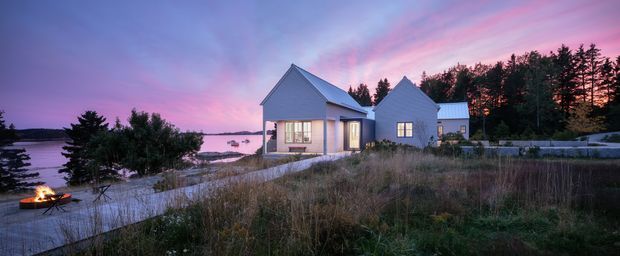 Exterior view of a contemporary coastal home at dusk, composed of several compact gabled volumes with pale cladding, large windows, and a low stone retaining wall. The house sits beside a calm waterfront with natural grasses, trees, and a small outdoor fire visible in the foreground.