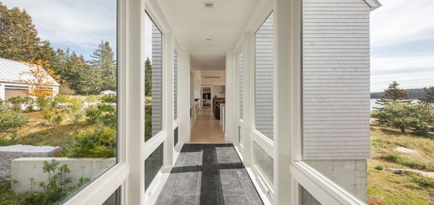A bright enclosed glazed hallway or entry corridor with large white-framed windows and doors on both sides, overlooking a landscaped coastal setting. The corridor has dark grey tiled flooring with a lighter central strip, white walls and trim, and leads toward an open interior living area. White horizontal weatherboard cladding is visible on the exterior, with water and trees beyond.