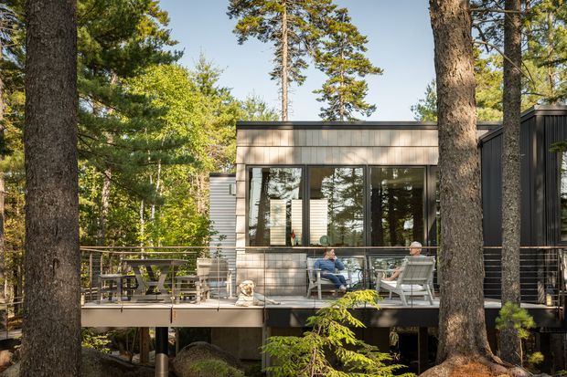 A modern elevated house nestled among mature trees in a wooded setting. The building has dark and natural-toned horizontal cladding, large reflective windows, and a broad timber deck with outdoor seating and dining furniture. The deck is supported above the forest floor, with railings and structural posts visible among the trees.