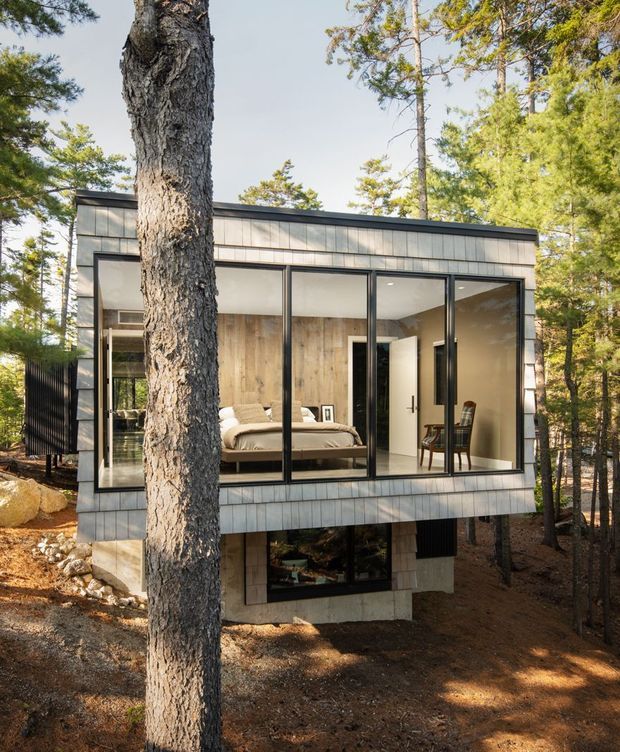 Exterior view of a compact modern cabin set among mature pine trees on a wooded, rocky site. The elevated two-storey structure has a box-like form, dark metal-framed floor-to-ceiling glazing, and grey shingle or panel cladding. A bedroom with a bed and chair is clearly visible through the upper-level glazing, while a partially visible lower-level room sits beneath the cantilevered upper volume.