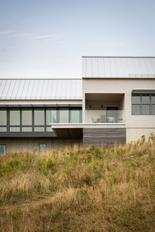 Exterior architectural photograph of a contemporary multi-level home set behind a dry grassy landscape. The building combines pale horizontal masonry, dark timber-like cladding, extensive dark-framed glazing, projecting volumes, and standing-seam metal roofs. A covered upper-level terrace with outdoor furniture is visible at the centre, while the lower level is partially concealed by the sloping vegetation.