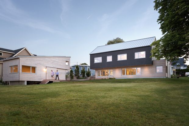 Exterior architectural view of a contemporary multi-building residential property across a broad lawn. The main two-storey building has a dark charcoal upper facade, light timber-toned lower cladding, a simple pitched metal roof, and regularly spaced rectangular windows. A smaller matching light-clad pavilion sits to the left, connected by a landscaped area and walkway. Mature trees and neighbouring homes frame the suburban setting.