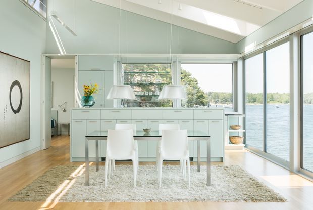 Bright open-plan dining and kitchen interior with floor-to-ceiling glazing overlooking a body of water. A pale mint-green island and cabinetry sit behind a glass dining table with white chairs, while timber flooring, a textured light rug, pendant lights, and abundant natural light create an airy, contemporary setting.