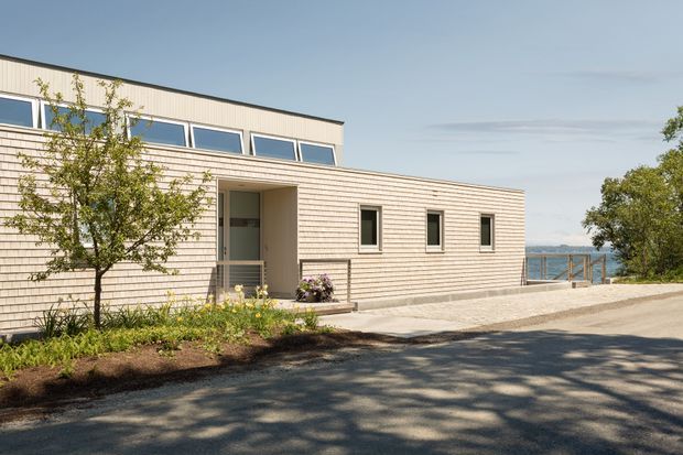 A low-rise modern coastal building with pale horizontal shingle-style cladding, a shallow low-pitched roof, clerestory windows, and a recessed glazed entry. The building sits beside a paved approach with simple landscaping, and water is visible beyond the side terrace and railing.