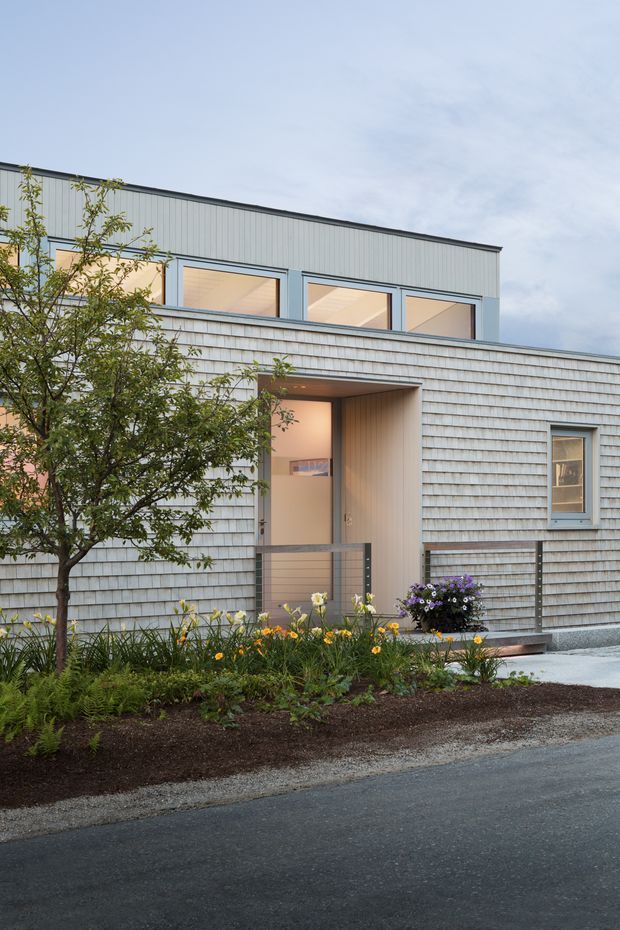 A contemporary low-rise residential building facade viewed from the street, featuring light grey shingle cladding on the lower level, vertically ribbed metal cladding above, large rectangular windows, and a recessed entry porch. A simple metal-and-glass railing borders the entrance ramp or steps, with a planted garden bed and small tree in the foreground.