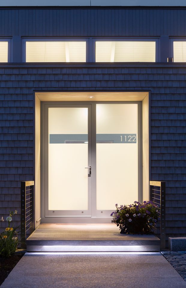 Night-time close-up of a contemporary residential entrance. A recessed double front door with frosted glazing and the number 1122 is framed by warm timber soffit lining and dark metal frames. The facade is clad in dark blue-grey shingle siding, with upper-level horizontal windows above. Timber decking, illuminated entry steps, dark cable railings, and potted flowering plants complete the welcoming entry.
