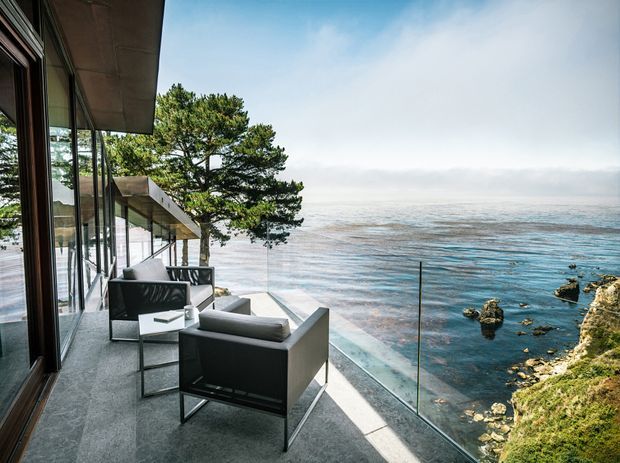 A modern coastal house terrace or balcony projects over a rocky ocean shoreline. The space features floor-to-ceiling glazing, dark-framed windows, a broad roof overhang, pale outdoor flooring, minimalist black-and-grey lounge chairs, and a frameless glass balustrade. A windswept coastal tree and clear blue ocean dominate the surrounding view.