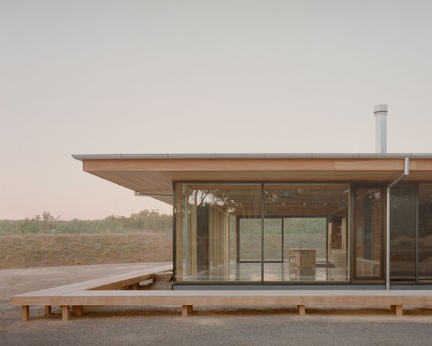 Single-storey modern pavilion-like residence set in a sparse rural landscape, photographed from outside. The building has a broad low-pitched roof with exposed timber soffit, extensive floor-to-ceiling glazing, dark slender window frames, and a raised timber deck wrapping the glazed living space. A simple metal flue projects above the roof. The interior is visible through the glass but the exterior architecture dominates.