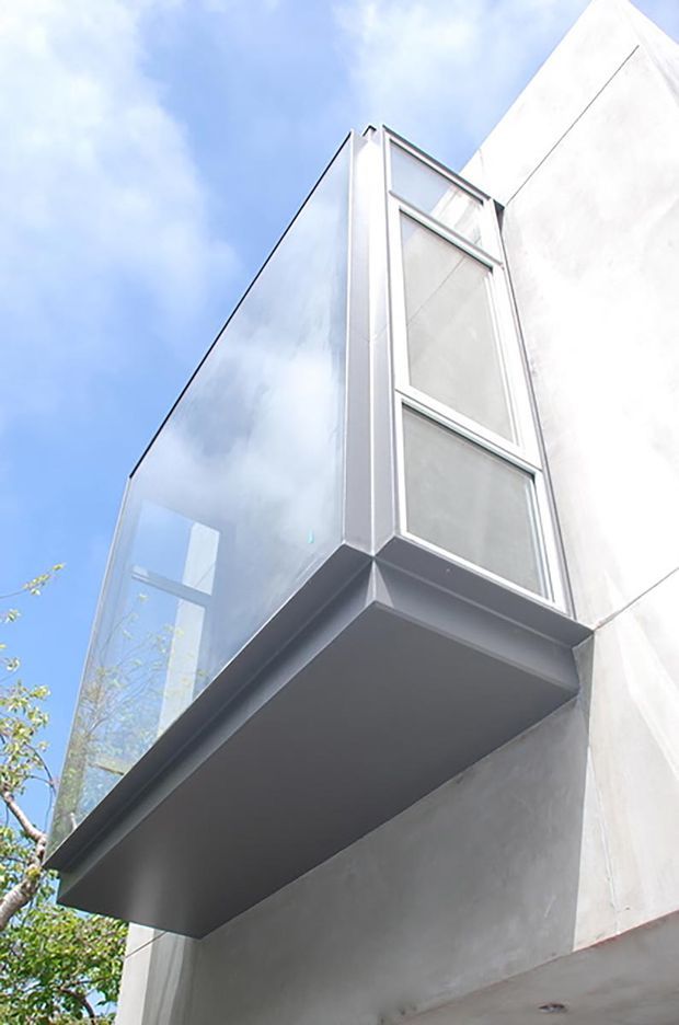 Upward-looking close-up of a minimalist modern building facade featuring a projecting cantilevered glazed volume or balcony. The structure has clear glass sides, slim light-coloured metal window frames, and smooth pale rendered or concrete walls, set against a bright blue sky.