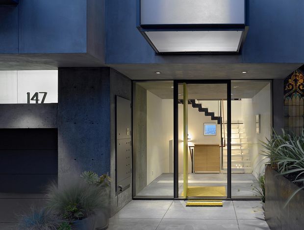 Street-facing entrance to a contemporary residence, featuring a dark blue and charcoal facade, large glazed entry doors, a recessed illuminated lobby, exposed stair structure, and integrated planter boxes. The number 147 is displayed beside a dark garage or service door. A stained-glass panel and indoor plants add decorative detail.